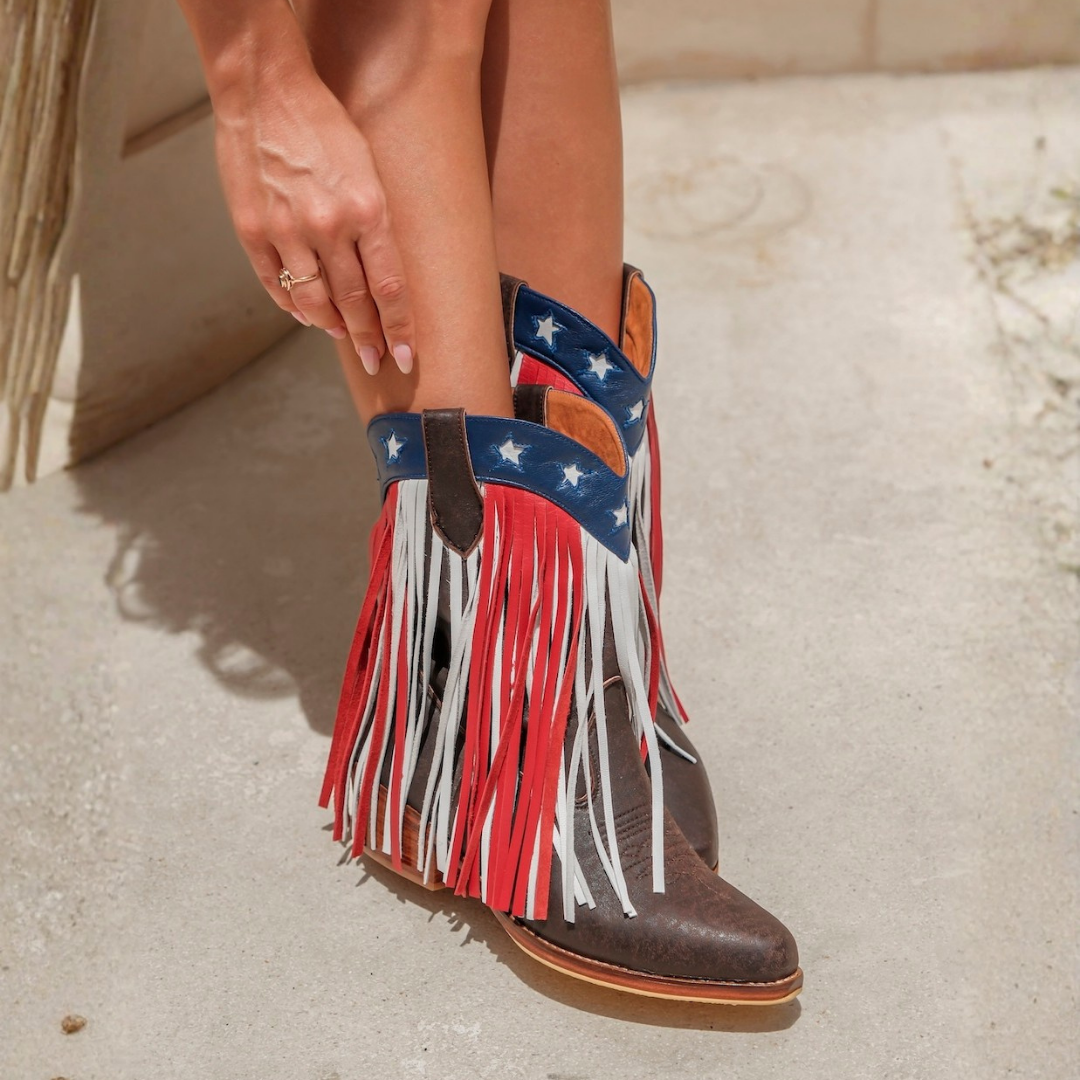 A close-up of a person wearing brown leather fringe cowboy boots with an American flag-inspired design. The boots feature red, white, and blue fringe, a blue strap with white stars, and a brown pointed toe, set against a light concrete background.