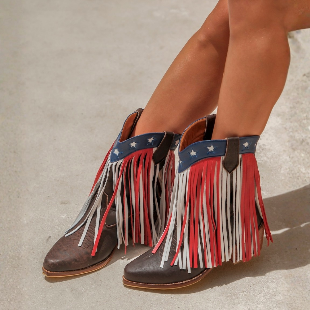 A close-up of a person's legs wearing brown leather fringe boots with a patriotic red, white, and blue design on the ankle strap and hanging tassels, set against a neutral concrete background.