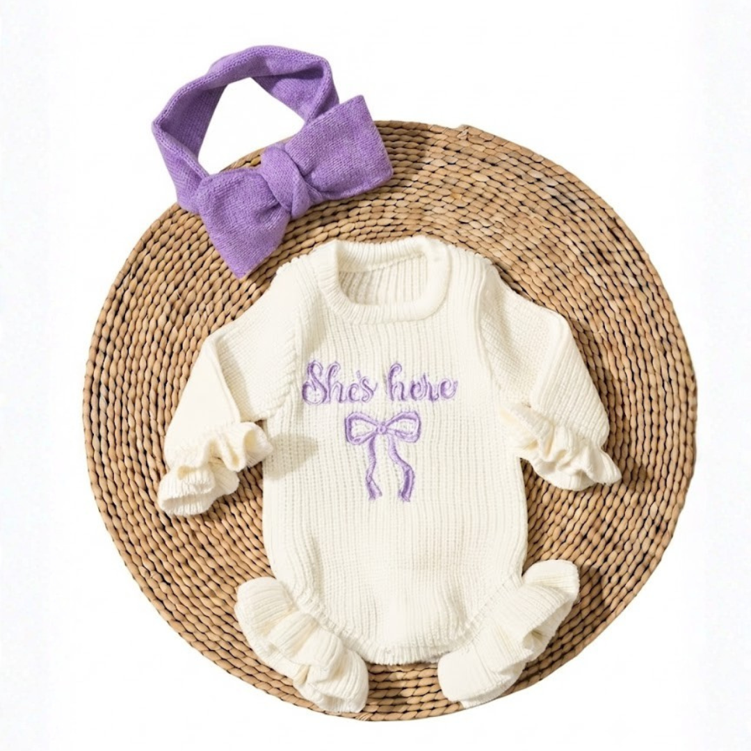 Little Darling Knit Romper Set from House of Hide in cream chunky ribbed knit with violet embroidered bow detail, ruffled cuffs and hem, button closure for easy diaper changes, matching violet bow headband being displayed on round woven mat