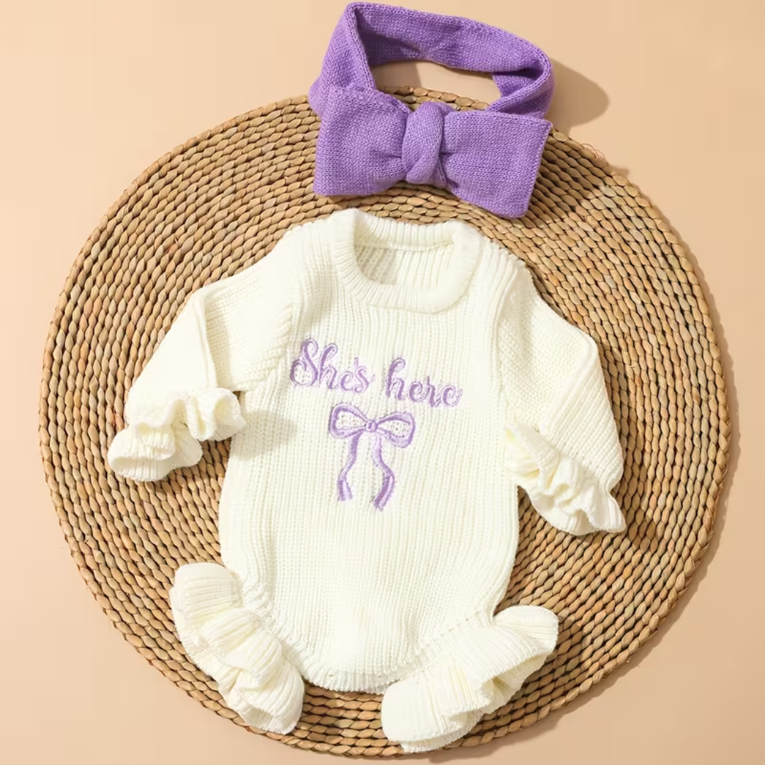 Little Darling Knit Romper Set from House of Hide in cream ribbed knit long-sleeve romper with ruffled edges, violet embroidered bow accent, coordinating violet bow headband being styled on round woven placemat with playful newborn aesthetic
