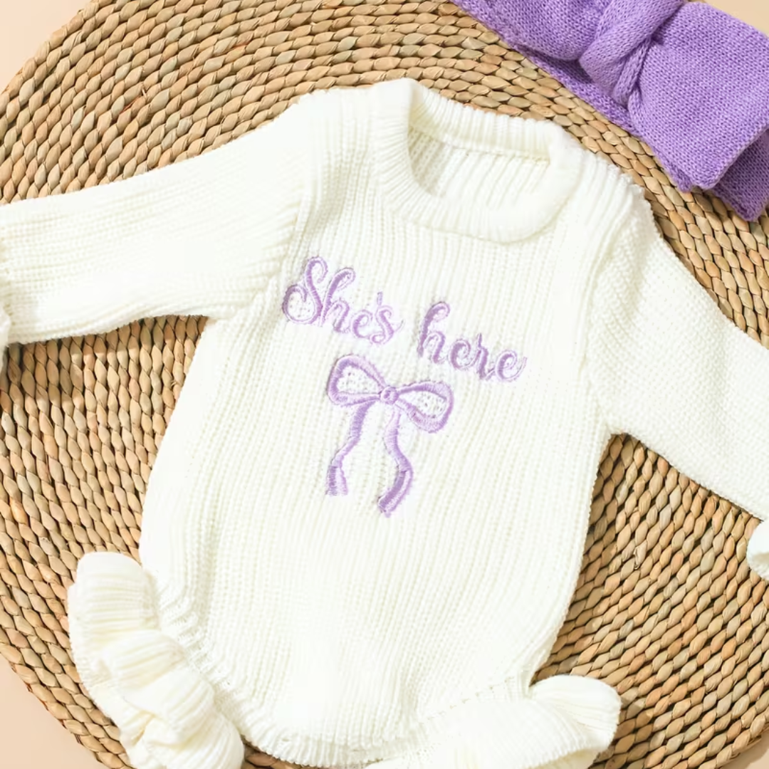 Little Darling Knit Romper Set from House of Hide in soft cream chunky ribbed knit baby romper with violet embroidered bow detail, button closure functionality, ruffled sweetness, coordinated violet bow headband being presented on round woven mat