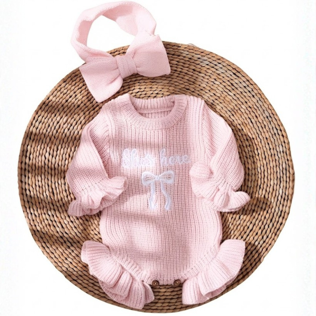 Little Darling Knit Romper Set from House of Hide in pale pink chunky ribbed knit with ruffled cuffs and hem, embroidered bow detail, matching pale pink bow headband being displayed on round woven mat with soft nursery aesthetic