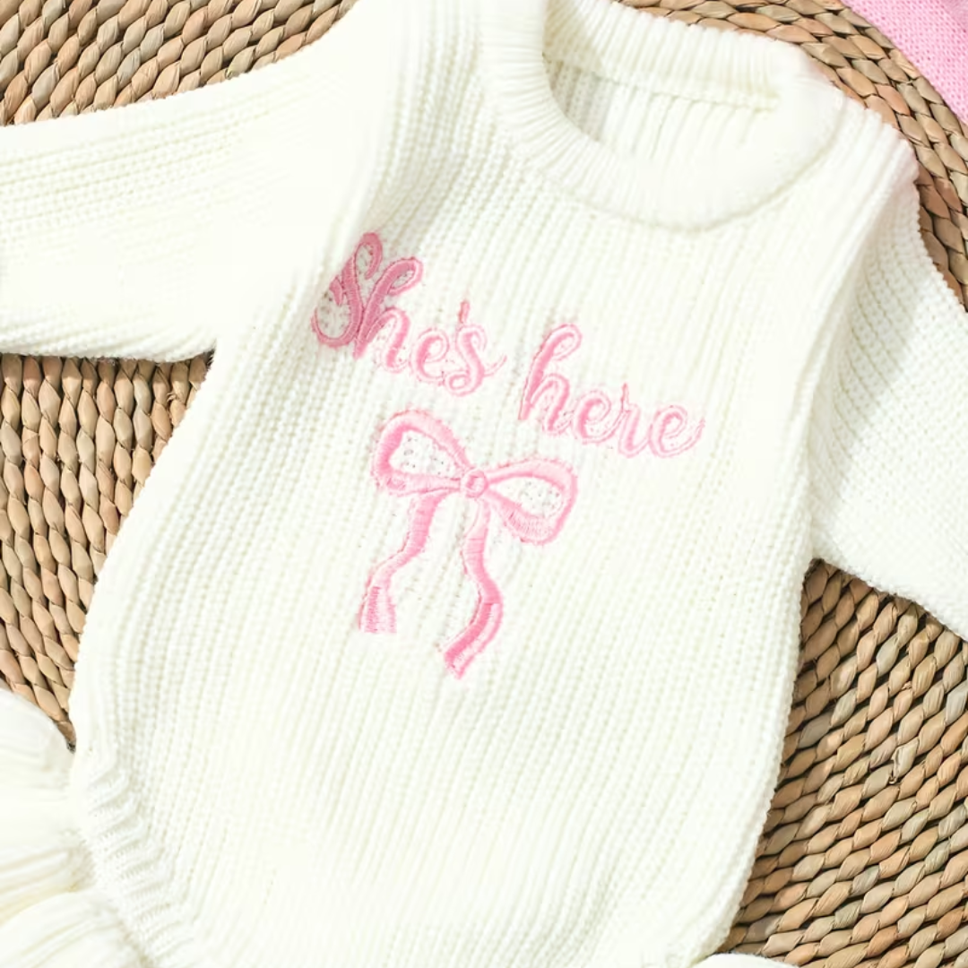 Little Darling Knit Romper Set from House of Hide in ivory chunky ribbed knit with pink embroidered she's here text and bow detail, coordinating pink bow headband being styled on round woven placemat backdrop