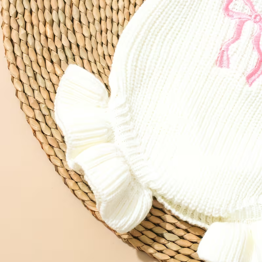 Little Darling Knit Romper Set from House of Hide in soft off-white ribbed knit romper with ruffled edges, pink embroidered bow graphic, matching pink bow headband accessory being displayed on woven circular mat