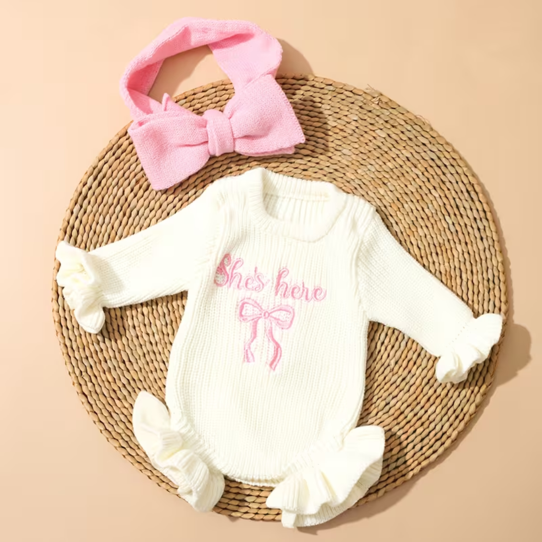 Little Darling Knit Romper Set from House of Hide in off-white chunky ribbed knit baby romper with pink embroidered announcement text and bow, matching pink bow headband being styled on round woven mat with neutral backdrop