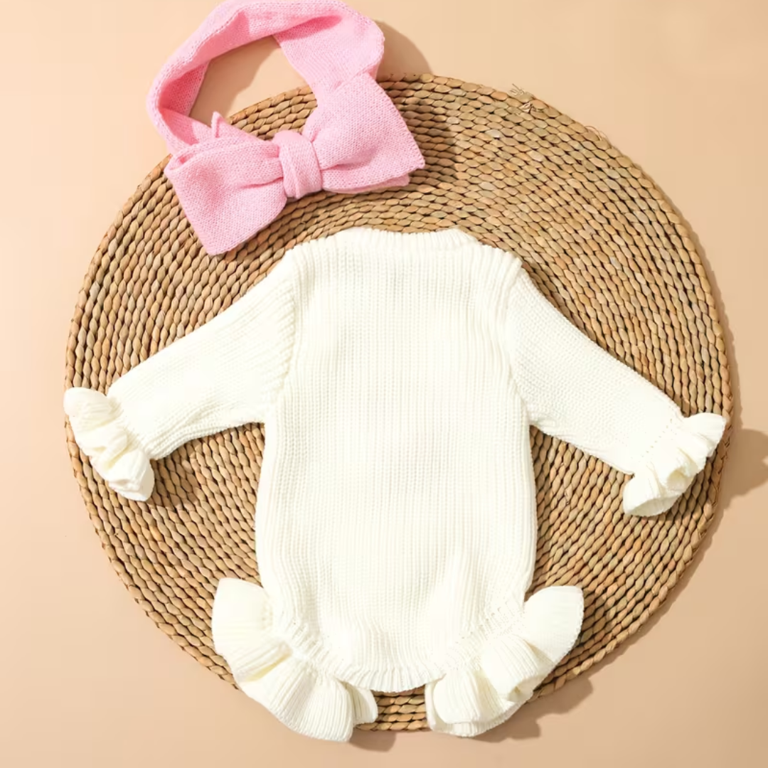 Little Darling Knit Romper Set from House of Hide in cream ribbed knit long-sleeve romper with ruffled trim, pink embroidered she's here announcement and bow, coordinating pink bow headband being displayed laid flat on round woven mat
