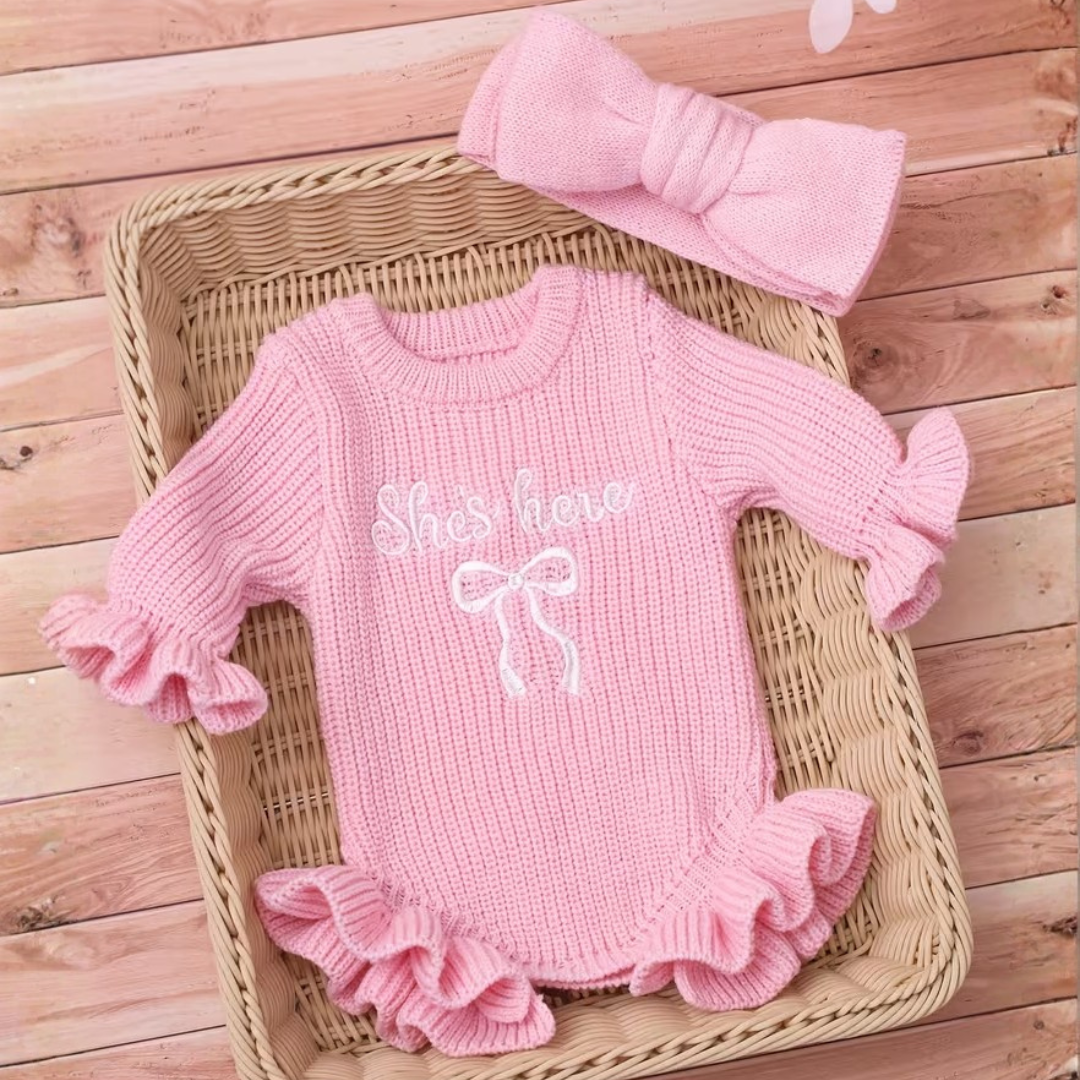 Little Darling Knit Romper Set from House of Hide in pink ribbed knit romper with ruffled sleeves and hem, white embroidered she's here text and bow design, matching pink bow headband being displayed in wicker basket on wooden plank background