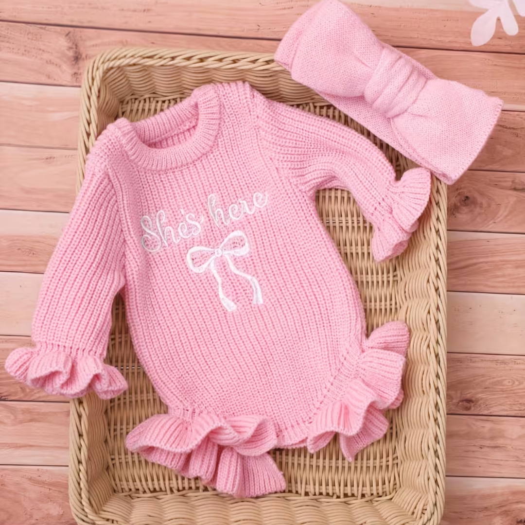 Little Darling Knit Romper Set from House of Hide in soft pink ribbed knit long-sleeve romper with ruffled edges and white embroidered bow, coordinated pink bow headband being displayed in wicker presentation on wooden surface