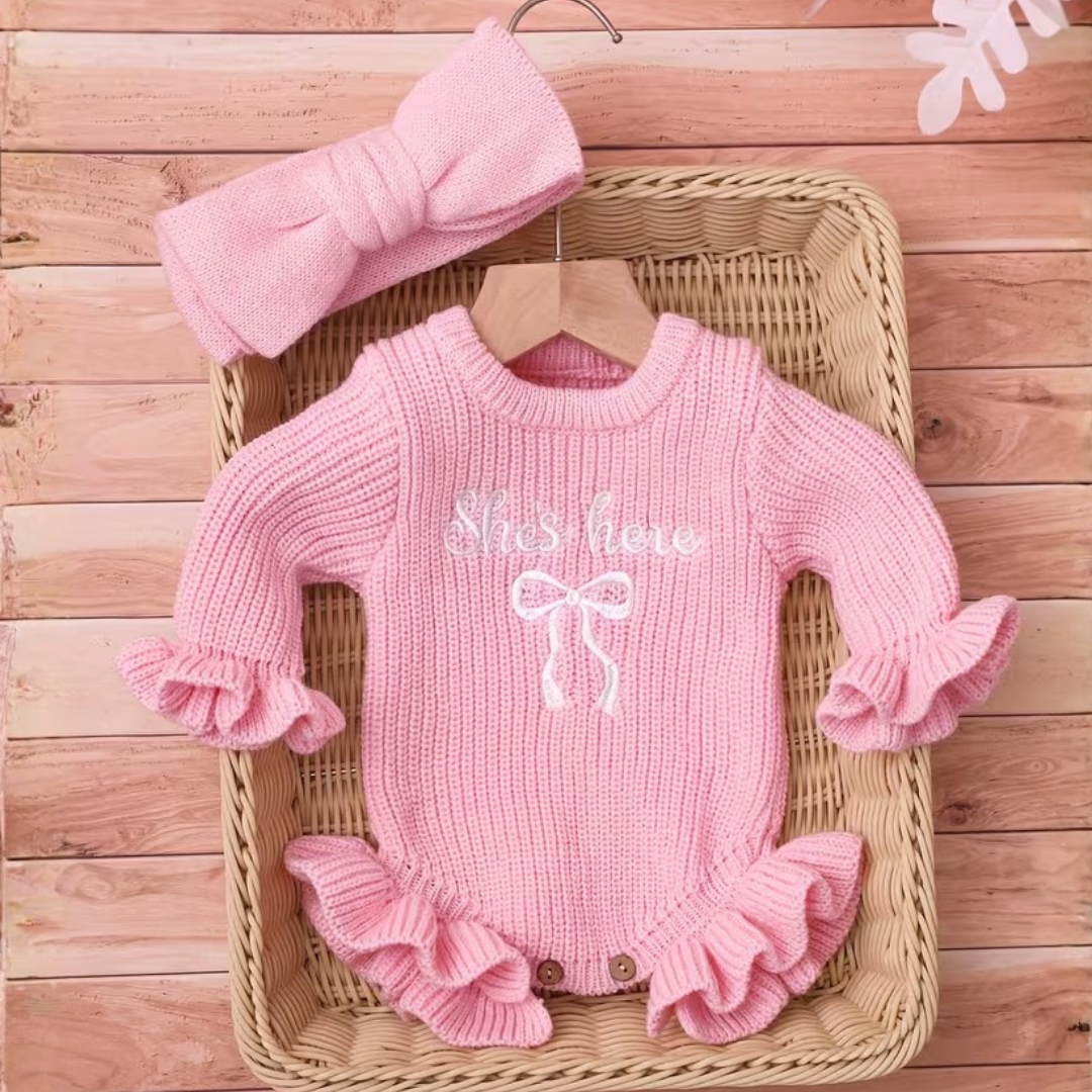 Little Darling Knit Romper Set from House of Hide in pink ribbed knit with white embroidered she's here announcement text, ruffled sleeves and hem, matching pink bow accessory being styled in wicker basket with rustic wood backdrop