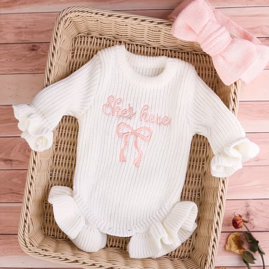 Little Darling Knit Romper Set from House of Hide in white ribbed knit with pink she's here embroidery and bow, ruffled edges, and matching pink pom-pom beanie being displayed in woven basket on pink wood flooring