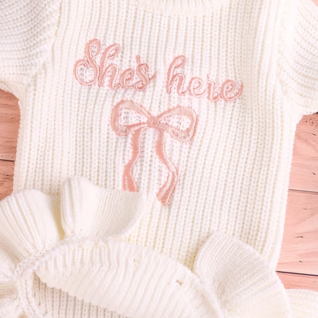 Little Darling Knit Romper Set from House of Hide in white ribbed knit romper with ruffled wrist and hem details, pink embroidered bow, and matching pink pom-pom beanie being displayed in editorial lifestyle setting