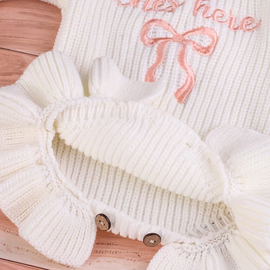 Close-up of Little Darling Knit Romper Set from House of Hide in white ribbed knit with pink embroidered she's here text and bow detail, ruffled edges, being displayed on woven basket with coordinating pink beanie