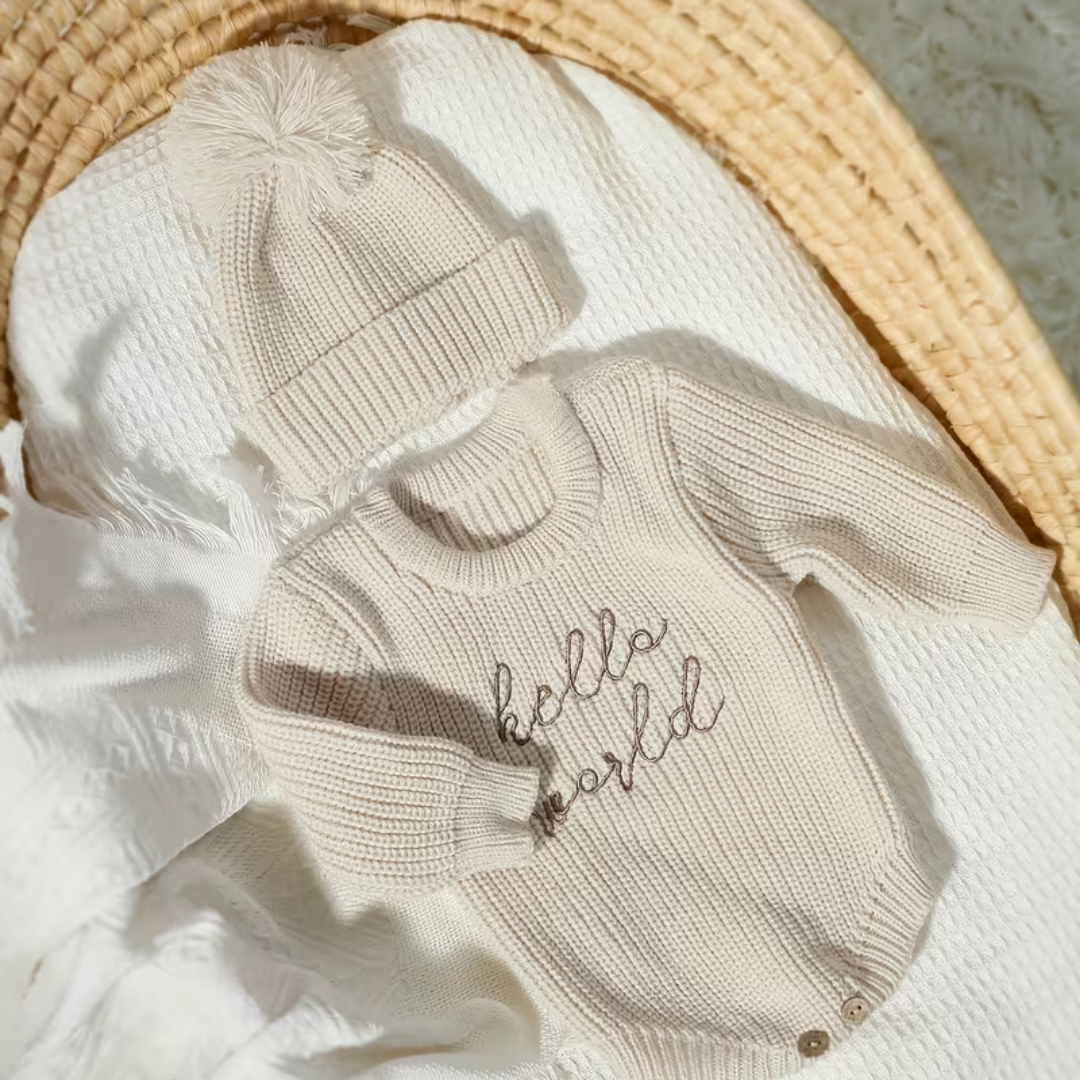 Little Wrangler Knit Romper Set from House of Hide in cream beige ribbed knit long-sleeve sweater with hello world embroidery and matching pom-pom beanie being displayed on white textured blanket inside woven basket