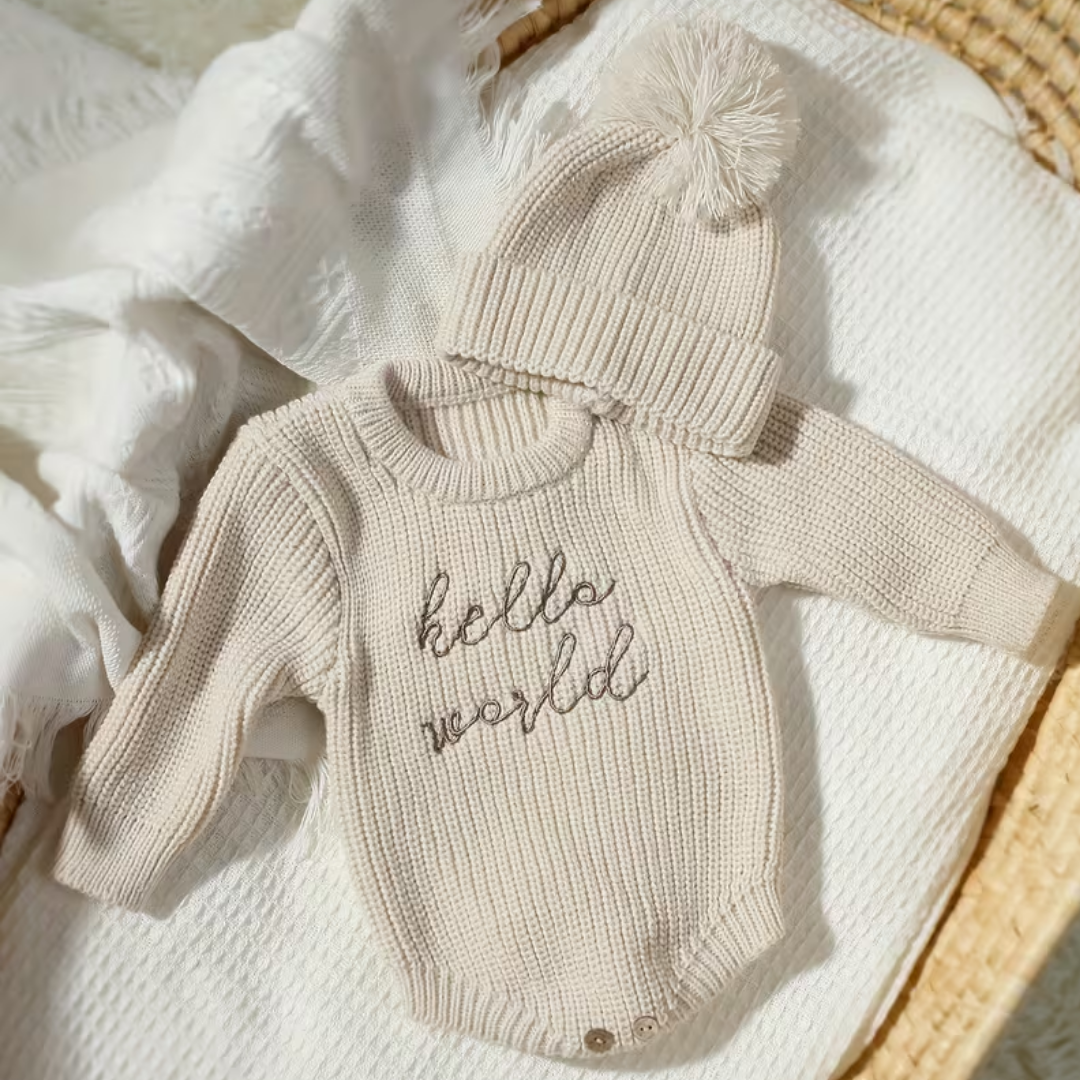 Little Wrangler Knit Romper Set from House of Hide in cream beige ribbed knit romper with dark script hello world embroidery and matching pom-pom beanie being displayed on textured white blanket