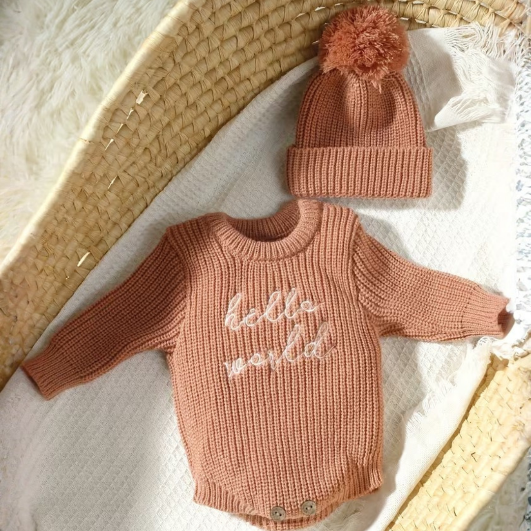 Little Wrangler Knit Romper Set from House of Hide in dusty terracotta pink ribbed knit long-sleeve bodysuit with white hello world text and matching pom-pom beanie being displayed on white textured blanket inside woven basket