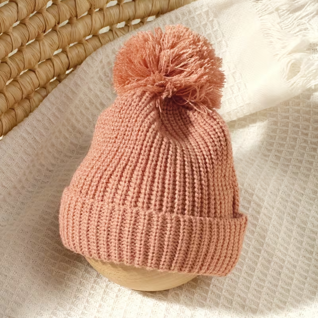 Little Wrangler Knit Romper Set from House of Hide in dusty pink ribbed knit pom-pom beanie being displayed on wooden stand over textured cream fabric with woven straw basket in background