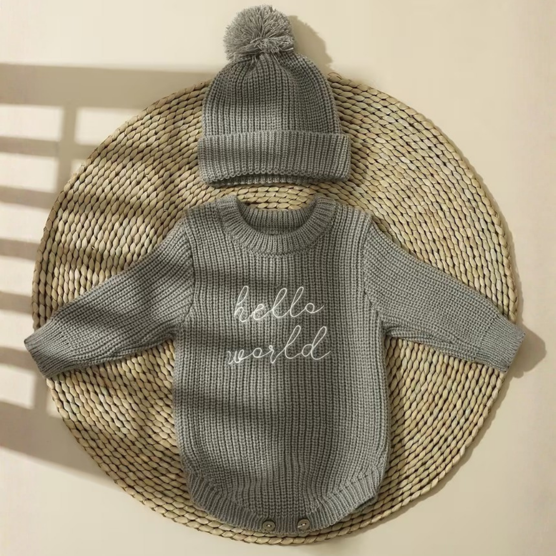 Little Wrangler Knit Romper Set from House of Hide in heather gray ribbed knit long-sleeve romper with white script hello world text and matching fluffy pom-pom beanie being displayed on round woven straw mat
