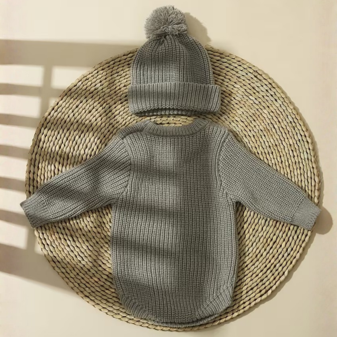 Little Wrangler Knit Romper Set from House of Hide in heather gray ribbed knit long-sleeve romper and matching pom-pom beanie being displayed on round woven straw mat with soft window shadow overlays