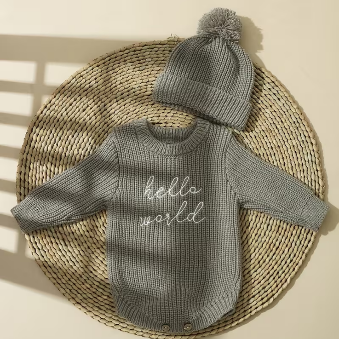 Little Wrangler Knit Romper Set from House of Hide in heather gray ribbed knit long-sleeve romper with white script hello world text and matching pom-pom beanie being displayed laid flat on round woven placemat