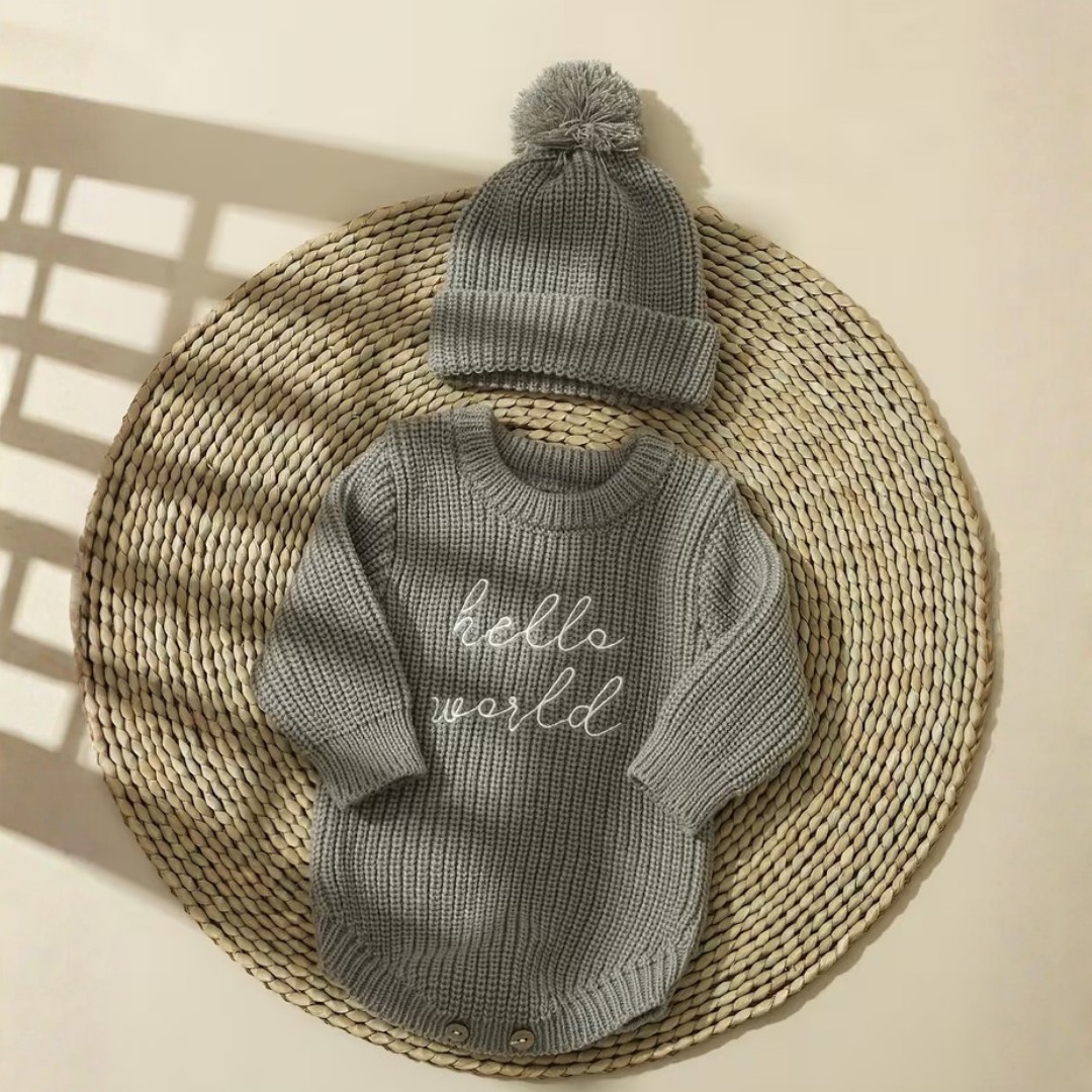 Little Wrangler Knit Romper Set from House of Hide in heather gray ribbed knit long-sleeve romper with white hello world text and matching pom-pom beanie being displayed on round woven placemat against warm beige background with soft window shadow overlays