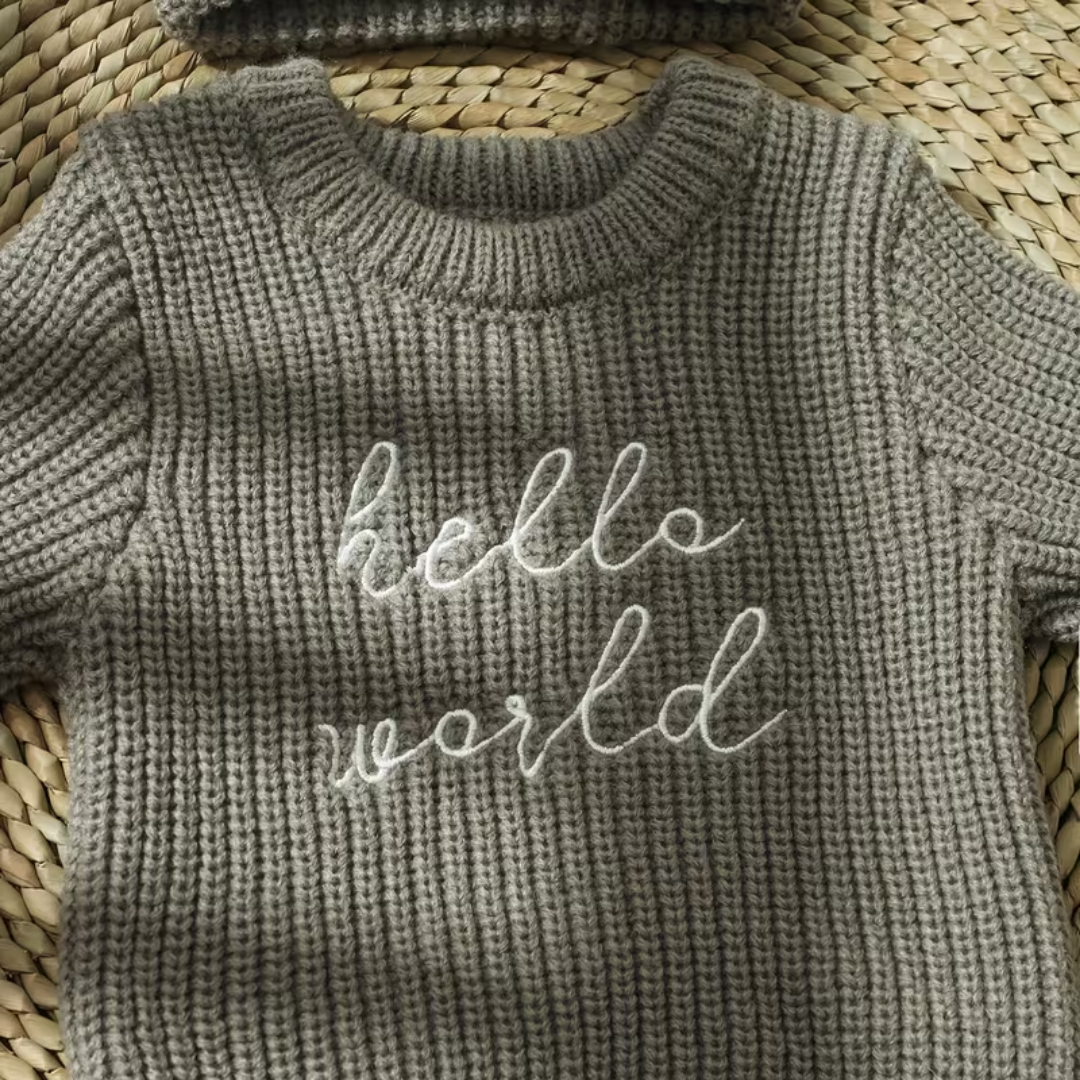 Little Wrangler Knit Romper Set from House of Hide in gray ribbed knit baby sweater with white script hello world embroidery being displayed in close-up on woven straw mat