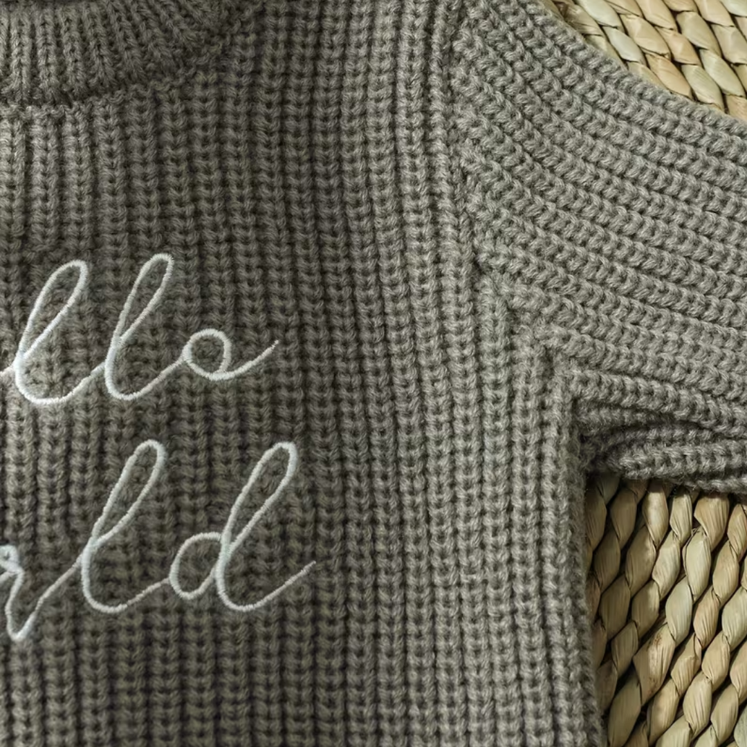 Little Wrangler Knit Romper Set from House of Hide in gray ribbed knit sweater with white cursive hello world embroidery being displayed in close-up on woven straw surface