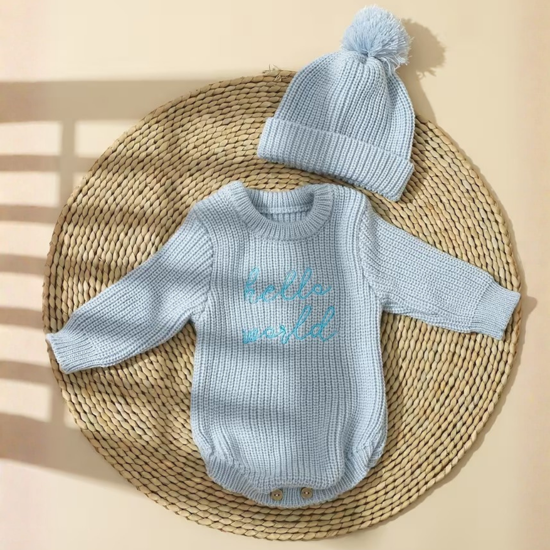 Little Wrangler Knit Romper Set from House of Hide in light blue ribbed knit long-sleeve bodysuit with hello world embroidery and matching pom-pom beanie being displayed on round woven mat