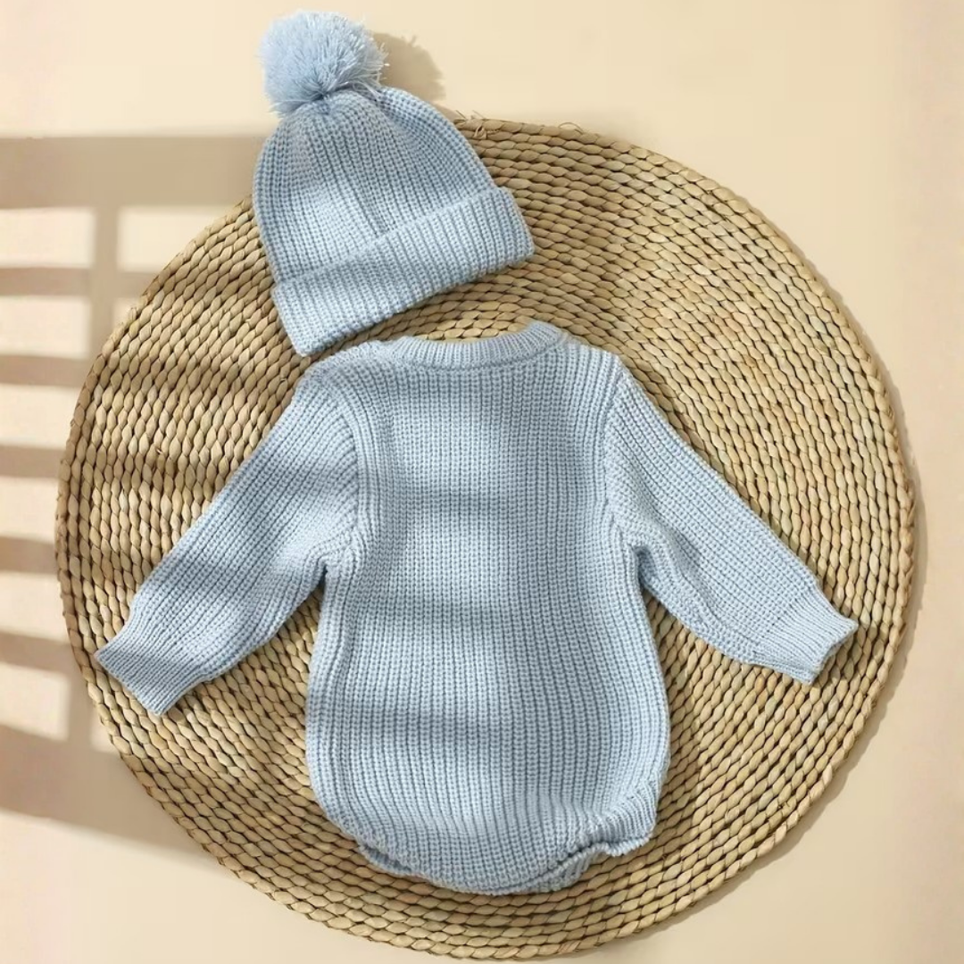 Little Wrangler Knit Romper Set from House of Hide in light blue ribbed knit long-sleeve romper and matching pom-pom beanie being displayed laid flat on round woven straw mat against warm beige background