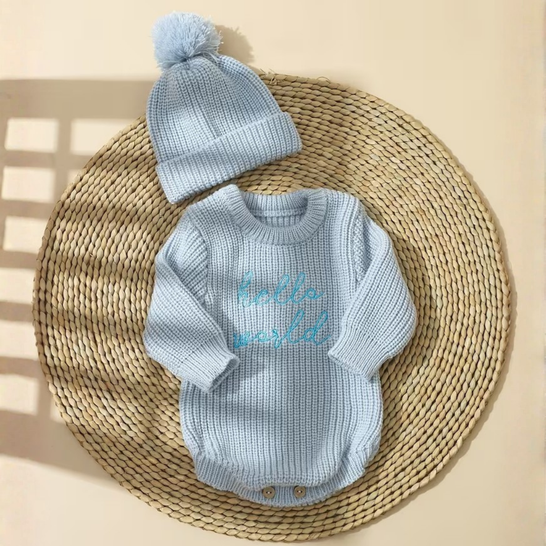 Little Wrangler Knit Romper Set from House of Hide in light blue ribbed knit long-sleeve bodysuit with hello world embroidery and matching pom-pom beanie being displayed on round woven mat