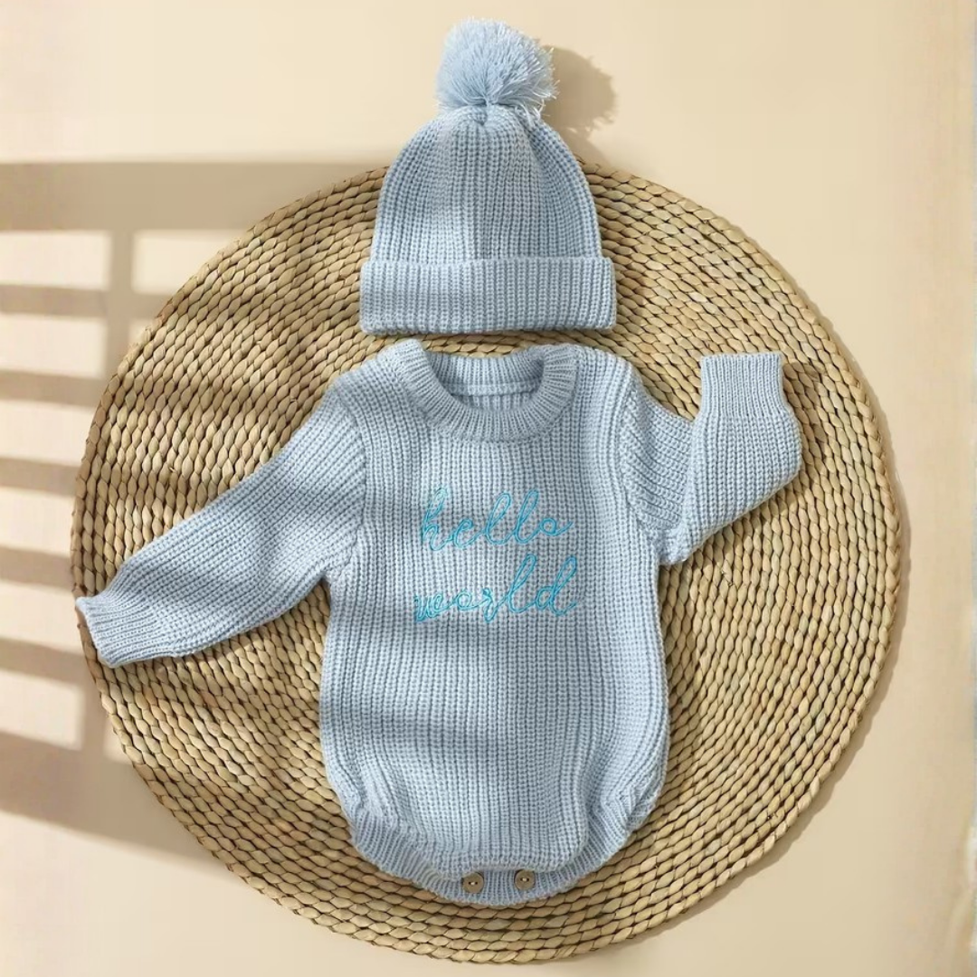 Little Wrangler Knit Romper Set from House of Hide in light blue ribbed knit long-sleeve romper and matching pom-pom beanie being displayed flat-lay on round woven placemat against warm beige background with soft window shadow overlays