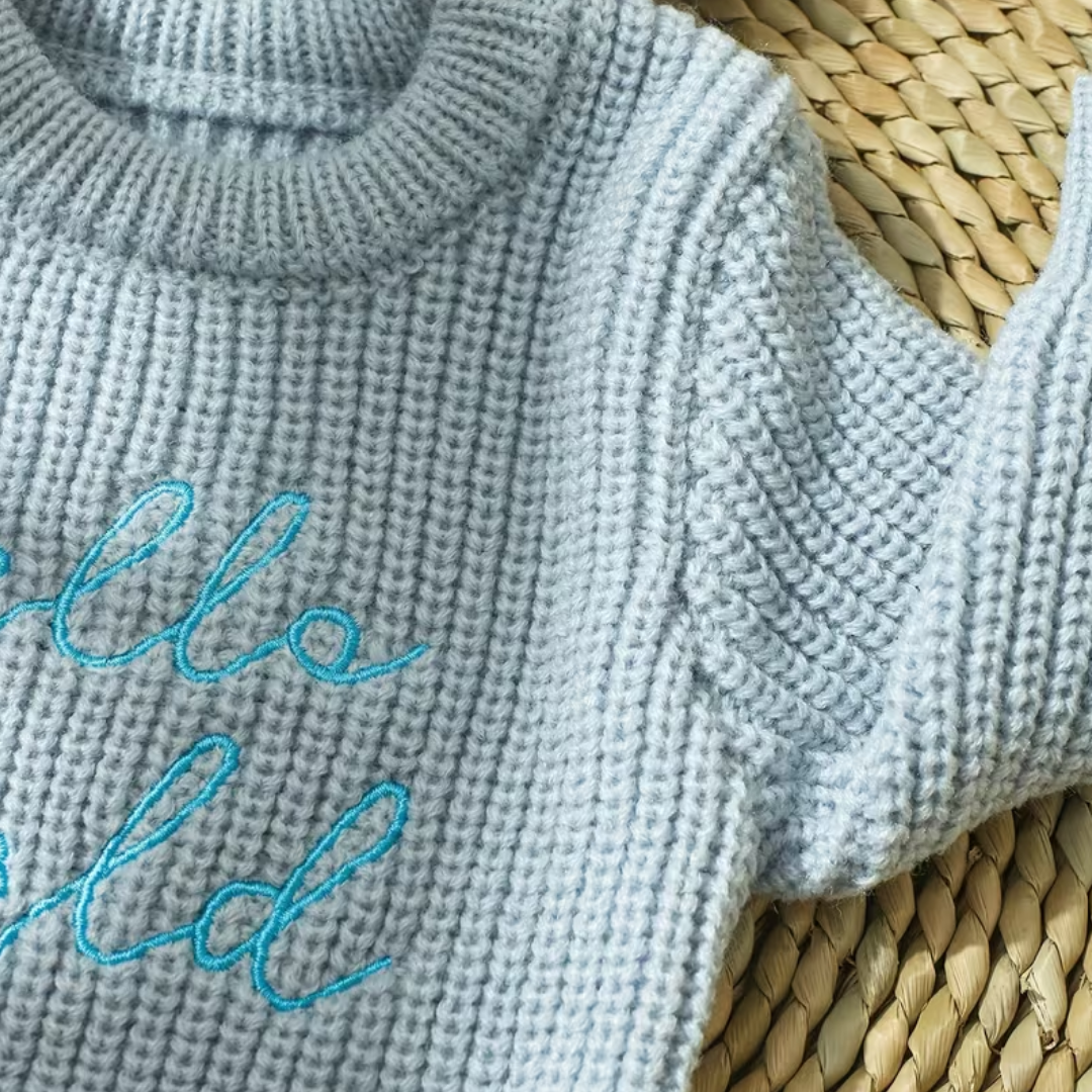Little Wrangler Knit Romper Set from House of Hide in light blue-gray ribbed knit sweater with bright turquoise embroidered script being displayed in close-up on woven straw surface