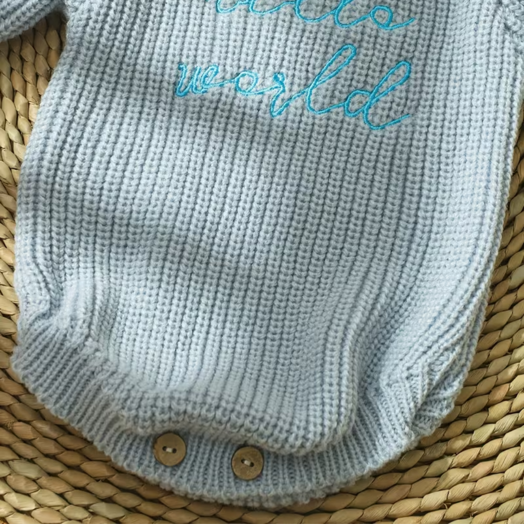 Little Wrangler Knit Romper Set from House of Hide in light gray ribbed knit baby bodysuit with turquoise embroidered the world text and wooden snap button closure being displayed in close-up on woven straw surface