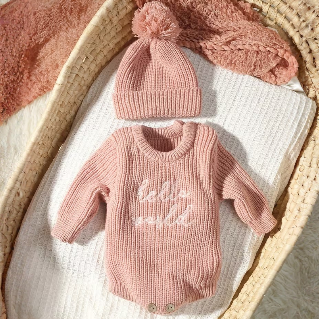 Little Wrangler Knit Romper Set from House of Hide in dusty pink ribbed knit long-sleeve romper with hello world text, matching pom-pom beanie, and fuzzy pink knitted hat being displayed in woven bassinet on white blanket