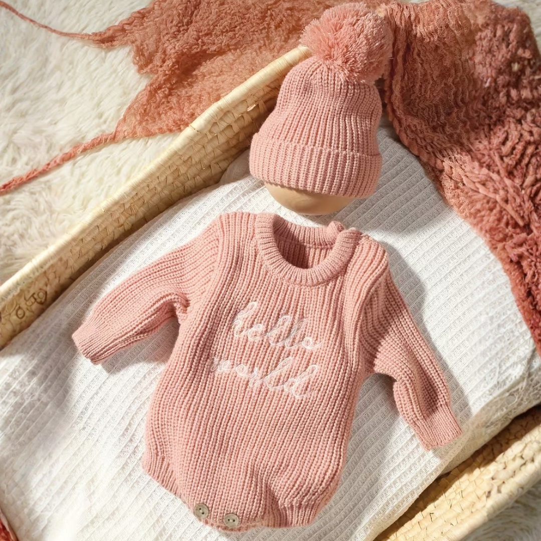Little Wrangler Knit Romper Set from House of Hide in dusty pink ribbed knit long-sleeve romper with hello world text and matching pom-pom beanie being displayed on white textured blanket inside woven basket with pink knit blanket