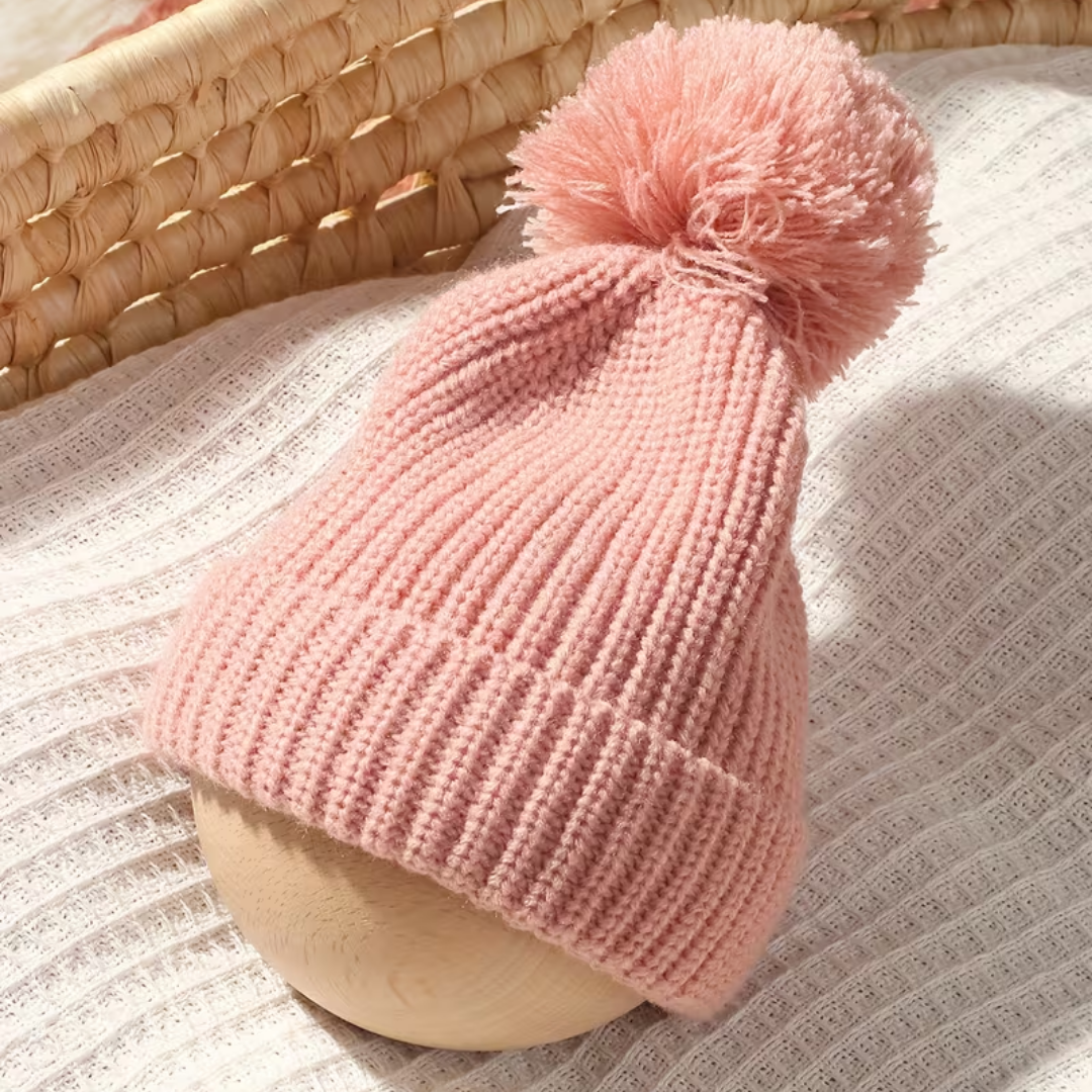 Little Wrangler Knit Romper Set from House of Hide in light pink ribbed knit beanie with fluffy pom-pom being displayed on wooden mannequin head over textured cream fabric with woven basket in background
