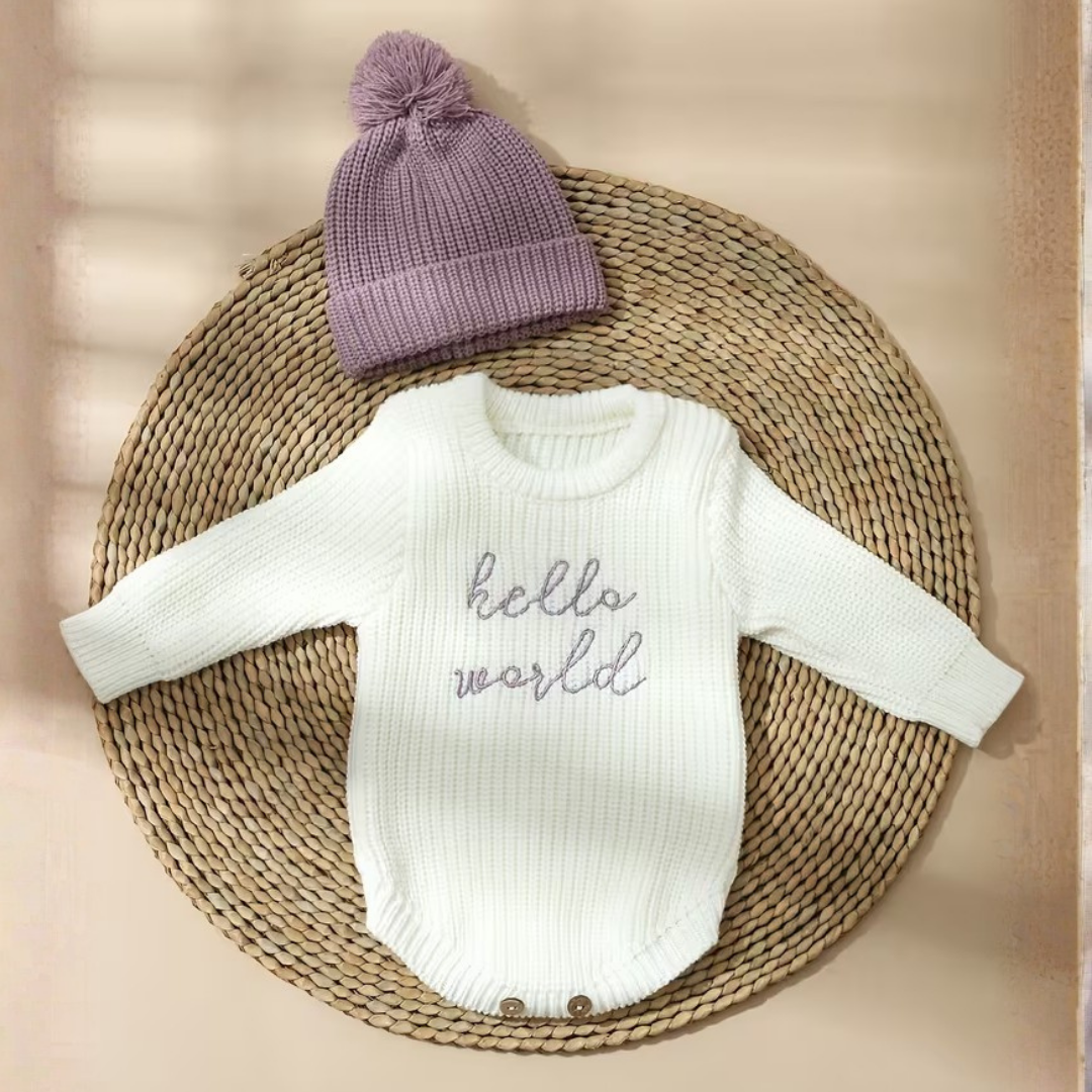 Little Wrangler Knit Romper Set from House of Hide in white ribbed knit with purple hello world text and lavender pom-pom beanie being displayed on round woven mat