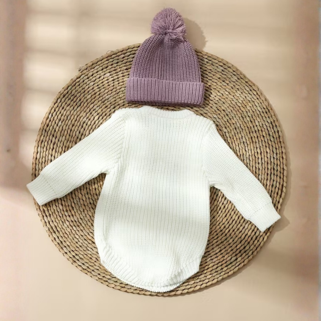 Little Wrangler Knit Romper Set from House of Hide in white ribbed knit bodysuit with dusty purple pom-pom beanie being displayed on round woven straw mat with soft window shadow overlays