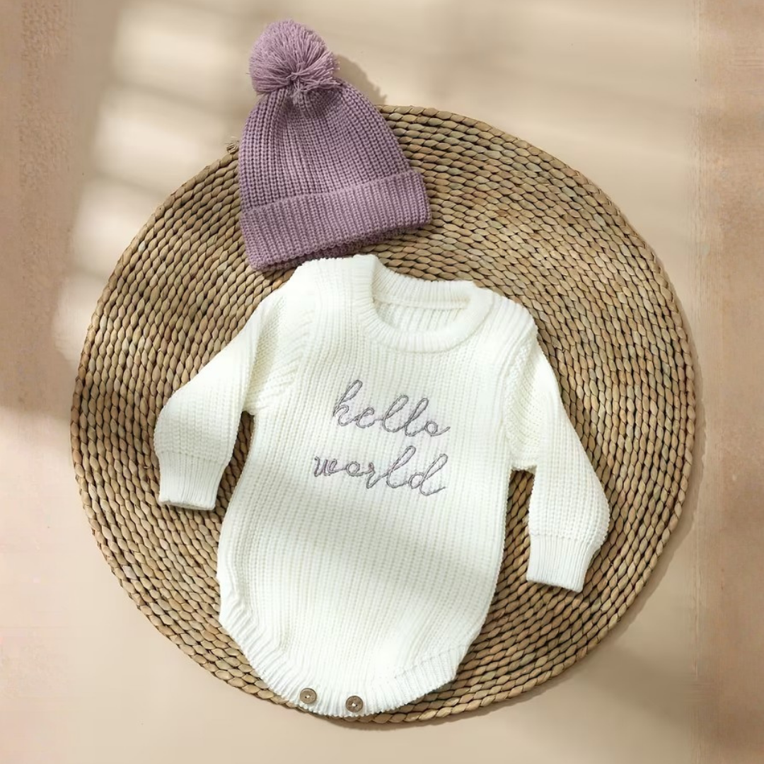 Little Wrangler Knit Romper Set from House of Hide in white ribbed knit with gray hello world text and lavender pom-pom beanie being displayed on round woven mat