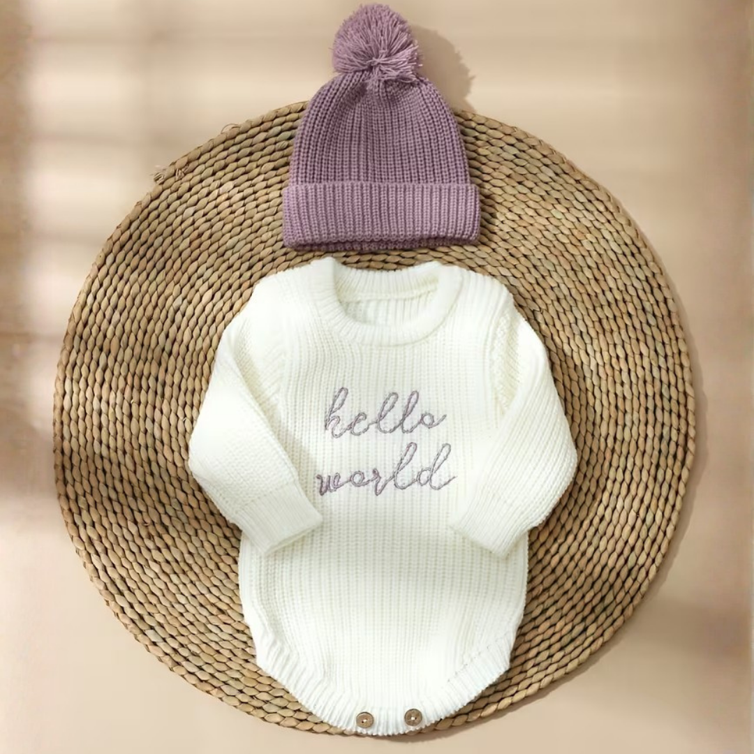 Little Wrangler Knit Romper Set from House of Hide in off-white ribbed knit with purple embroidered hello world text and dusty lavender-purple pom-pom beanie being displayed on round woven mat