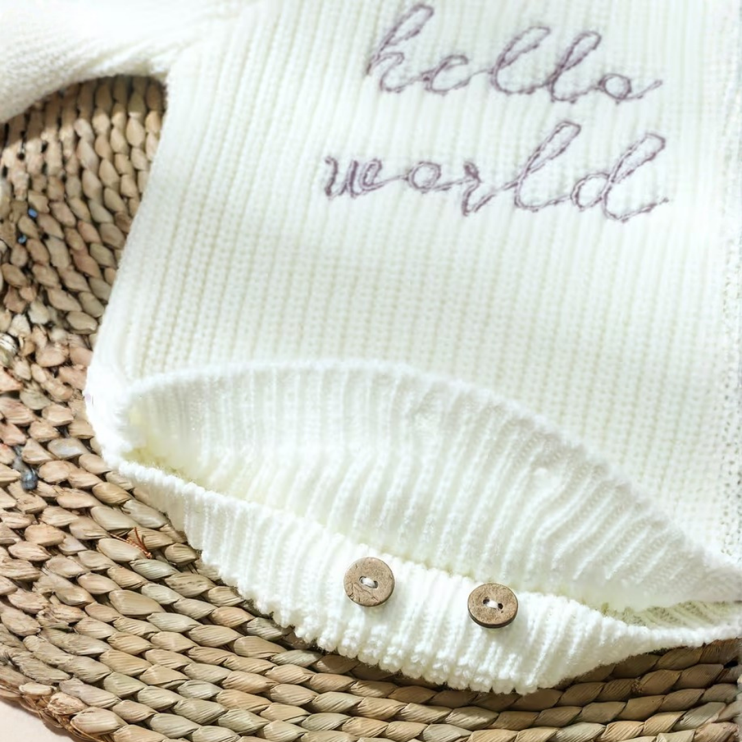 Little Wrangler Knit Romper Set from House of Hide in off-white ribbed knit with hello world embroidered text, wooden crotch buttons, being displayed on woven straw mat