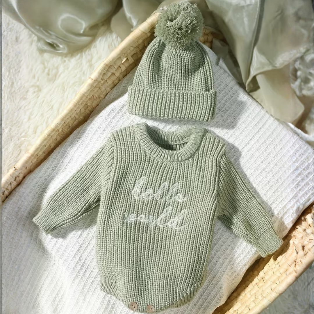 Little Wrangler Knit Romper Set from House of Hide in soft sage green ribbed knit long-sleeve romper with hello world embroidery and matching pom-pom beanie being displayed on white textured blanket inside woven basket