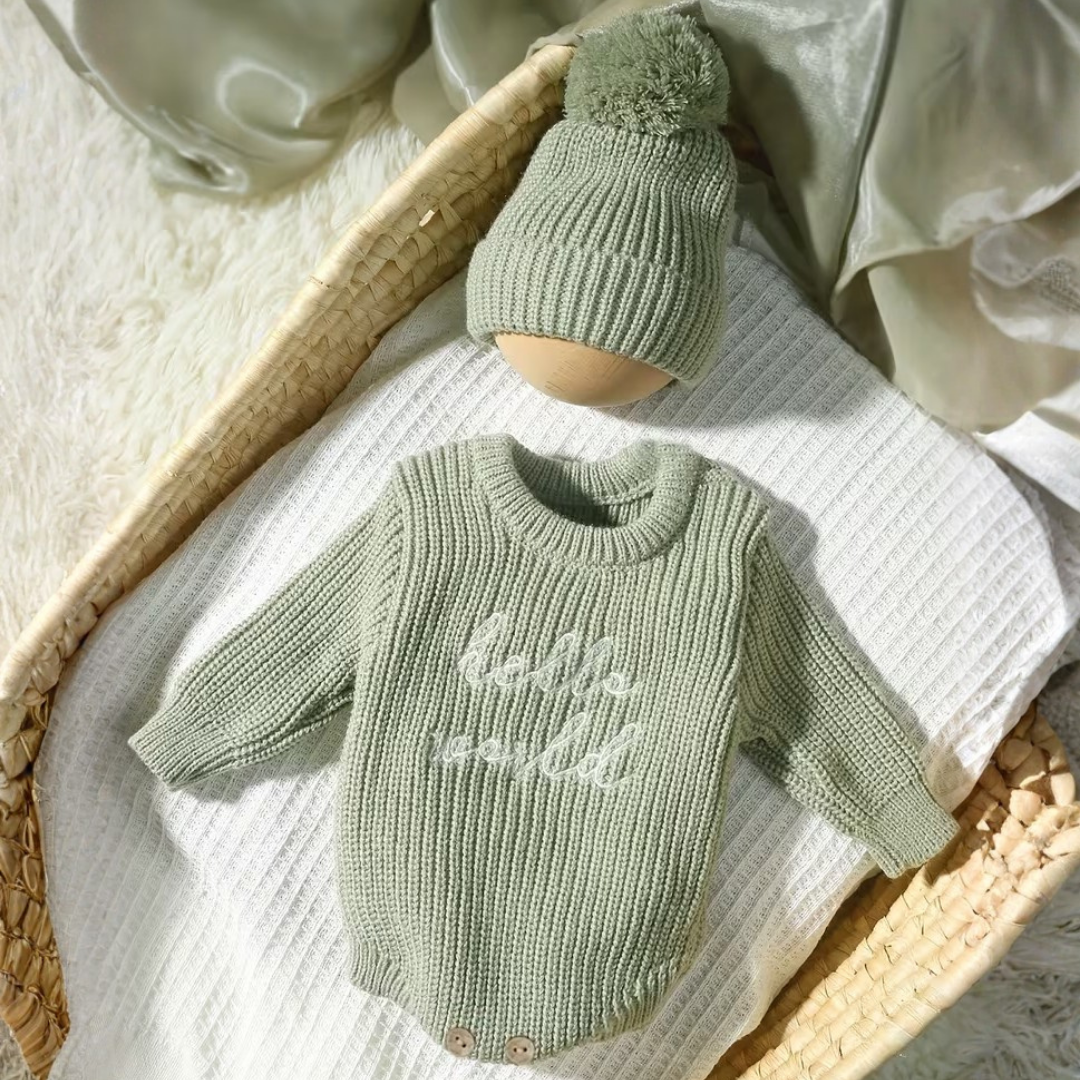 Little Wrangler Knit Romper Set from House of Hide in sage green ribbed knit long-sleeve bodysuit with hello world embroidery and matching pom-pom beanie being displayed in woven basket