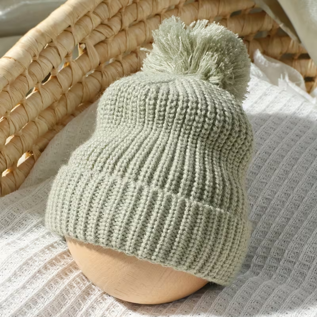 Little Wrangler Knit Romper Set from House of Hide in sage green ribbed knit beanie with pom-pom being displayed on mannequin head against wicker chair and textured fabric background
