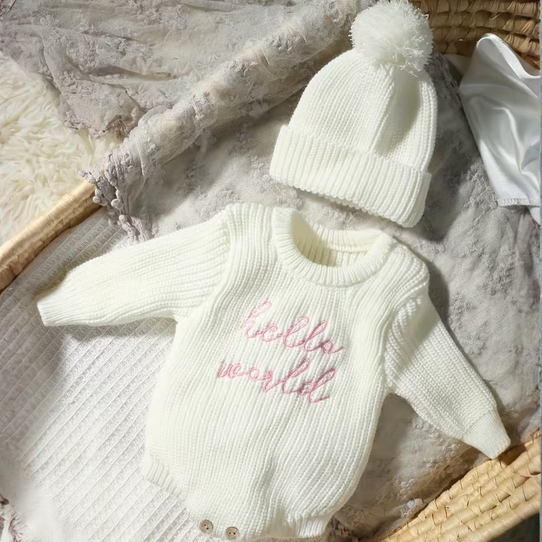 Little Wrangler Knit Romper Set from House of Hide in cream beige ribbed knit long-sleeve bodysuit with pink script hello world embroidery and matching pom-pom beanie being displayed in wicker basket with soft blankets