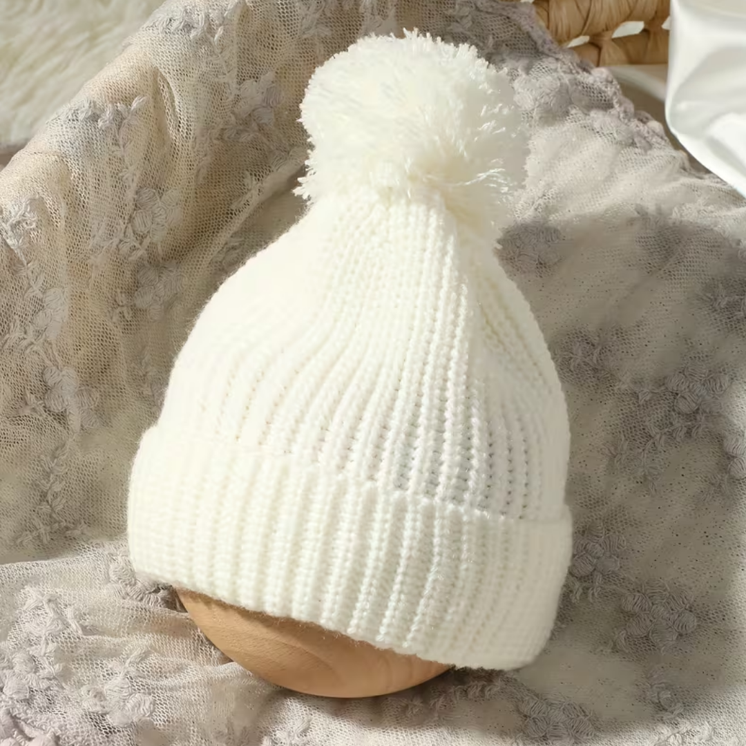 Little Wrangler Knit Romper Set from House of Hide in cream beige ribbed knit beanie with fluffy pom-pom being displayed on wooden stand against soft beige lace fabric background