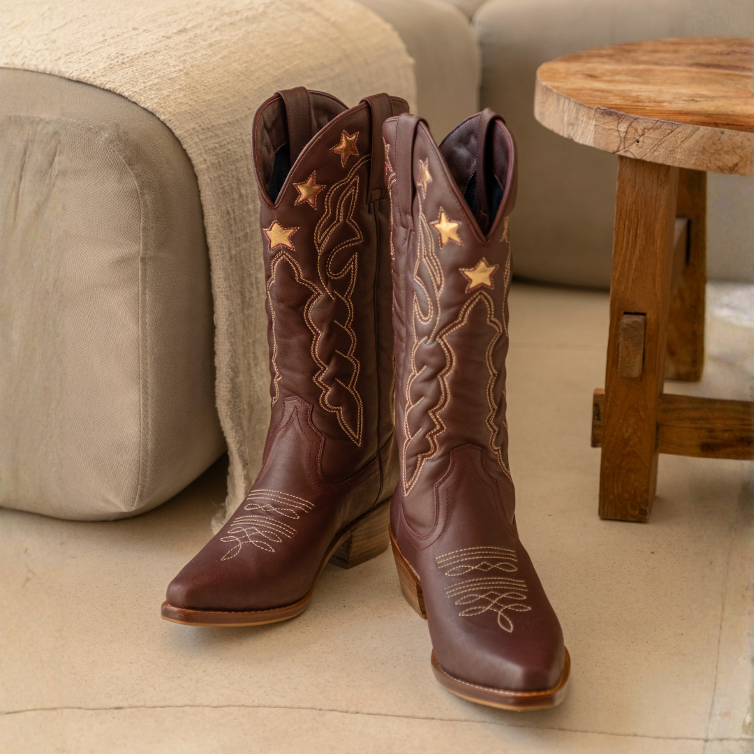 Lone Star Boots from House of Hide in dark brown leather with gold embroidered star and scroll designs being displayed standing on light beige tiled floor in living room setting with sofa and wooden stool