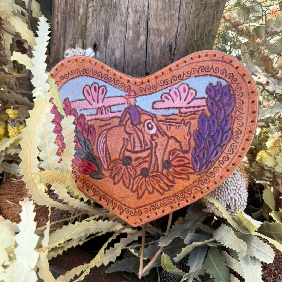 A heart-shaped handcrafted wooden plaque featuring a western cowboy and horse design, set against a rustic outdoor background with leaves and a wooden post.