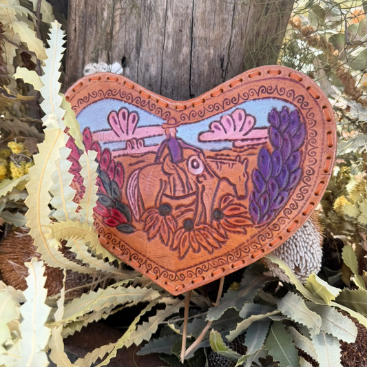 A heart-shaped handcrafted wooden plaque featuring a western cowboy and horse design, set against a rustic outdoor background with leaves and a wooden post.