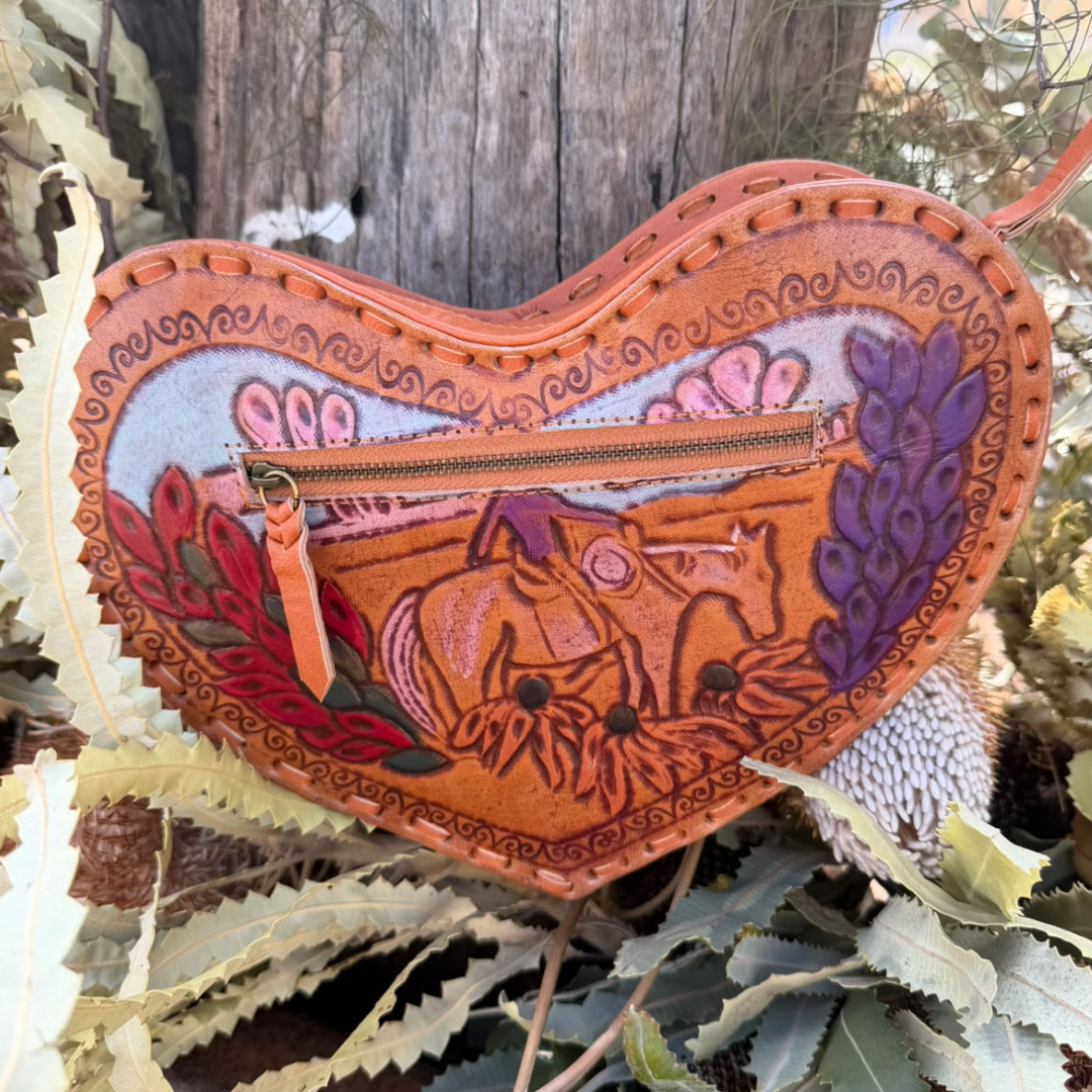 A heart-shaped hand-tooled leather clutch or wallet featuring a detailed western motif with a horse, purple foliage, and red leaves, set against a rustic outdoor background of leaves and a wooden post.