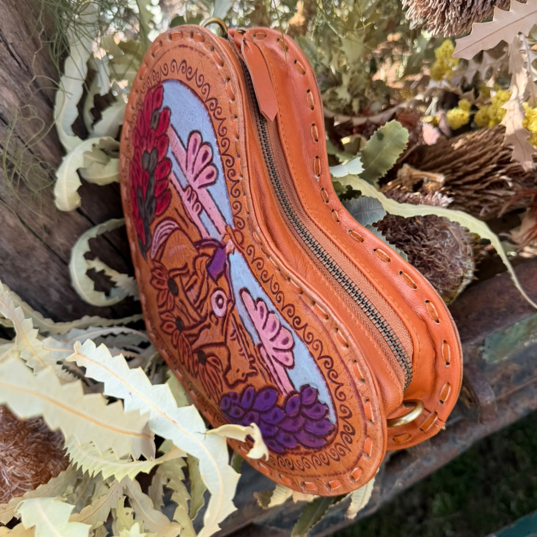 A handcrafted tan leather pouch with a colorful hand-painted design featuring a deer, pink flowers, and purple grapes, styled outdoors among dried leaves and natural elements.